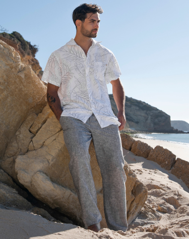 Homme contre une roche sur la plage portant une chemise fleurie gris et blanc et un pantalon gris de la marque Off the Beach.