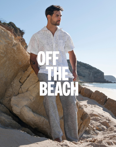 Homme contre une roche sur la plage portant une chemise fleurie gris et blanc et un pantalon gris de la marque Off the Beach.