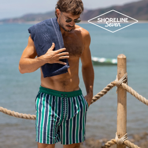 A man holding his towel by the water and a fence, wearing green, blue, and white striped swim trunks from the brand Shoreline Seven.