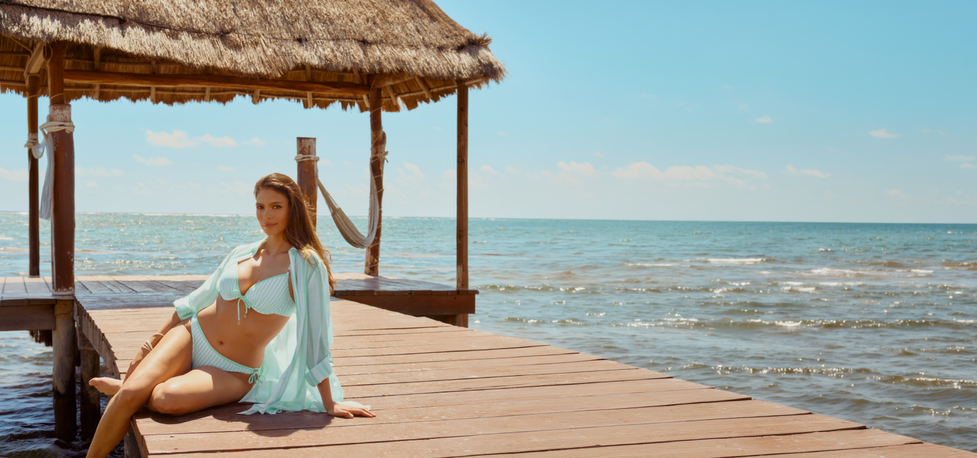 Femme assise sur un quai au bord de l'eau portant un ensemble coordonn&eacute; d'un ensemble bikini et chemise assortie lign&eacute; turquoise et blanc de la marque Turquoise Couture.