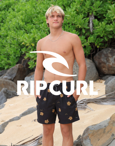A man standing on rocky sand, wearing floral-print shorts from the Rip Curl brand.