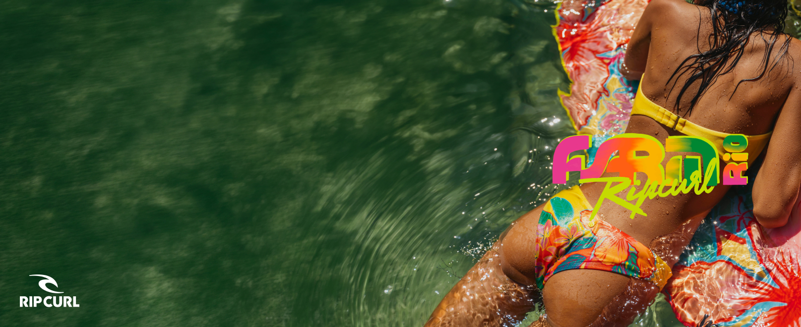 A woman in the water, viewed from behind and partially lying on a surfboard, wearing a bikini set from the Rip Curl x FARM Rio collaboration.
