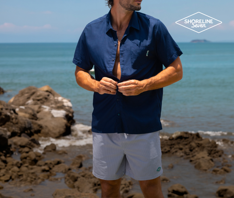A man standing by the water, buttoning up his navy blue shirt to complete his striped shorts outfit from the Shoreline Seven brand.