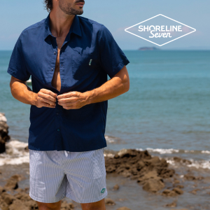 A man standing by the water, buttoning up his navy blue shirt to complete his striped shorts outfit from the Shoreline Seven brand.