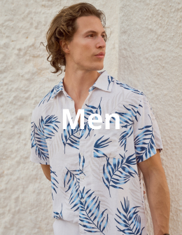 A man standing against a wall, wearing a palm tree print shirt and white pants from the Off The Beach brand.	
