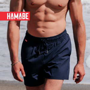 A man walking along the beach, wearing blue swim trunks from Hamabe.