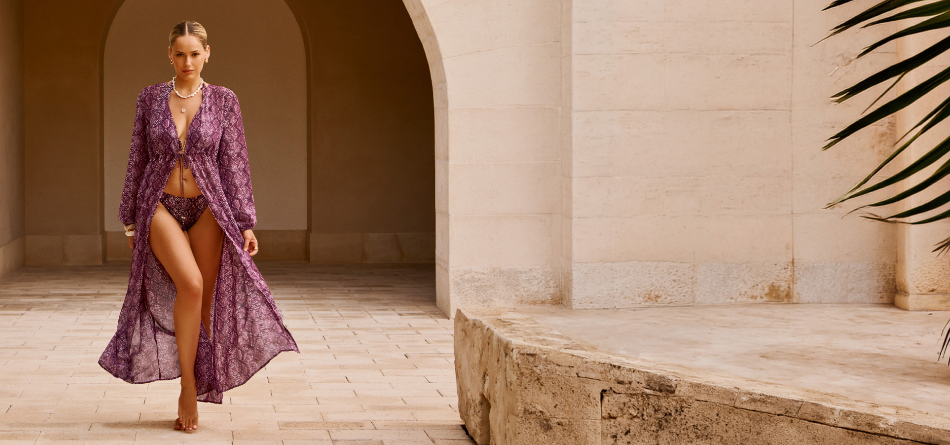 A woman walking through an architectural building, wearing a purple snakeskin-print bikini set and its matching kimono from the Santemare brand.