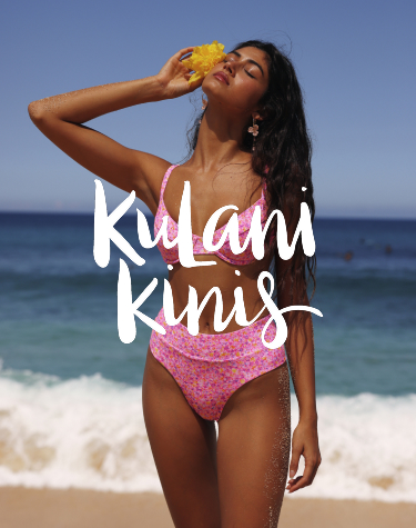 Femme marchant sur la plage, v&ecirc;tue d&rsquo;un bikini rose &agrave; imprim&eacute; floral de la marque Kulani Kinis, avec une fleur d&eacute;licatement gliss&eacute;e dans ses cheveux, pour une allure tropicale.