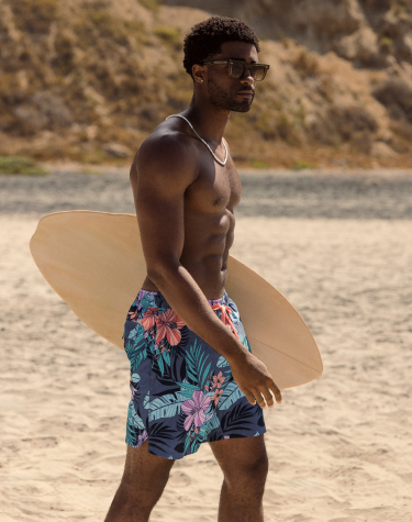Homme debout sur la plage tenant une petite planche de surf, vêtu d’un maillot avec un imprimé de feuillage tropical dans des tons bleus et roses éclatants, prêt pour une aventure au bord de la mer.