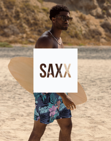 Homme debout sur la plage tenant une petite planche de surf, v&ecirc;tu d&rsquo;un maillot avec un imprim&eacute; de feuillage tropical dans des tons bleus et roses &eacute;clatants, pr&ecirc;t pour une aventure au bord de la mer.