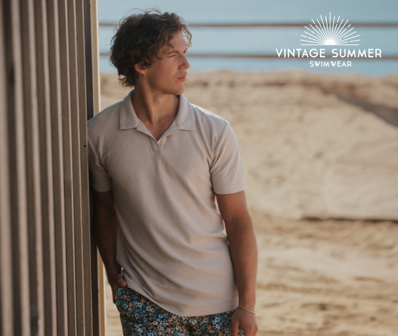 Homme debout sur la plage, v&ecirc;tu d&rsquo;un t-shirt beige et d&rsquo;un short de bain, profitant d&rsquo;un moment d&eacute;contract&eacute; au bord de la mer.