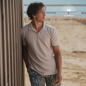 Homme debout sur la plage, vêtu d’un t-shirt beige et d’un short de bain, profitant d’un moment décontracté au bord de la mer.