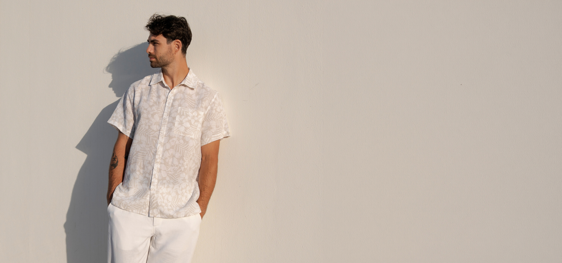 A man leaning against a wall, wearing a floral-patterned linen shirt and white linen pants. 