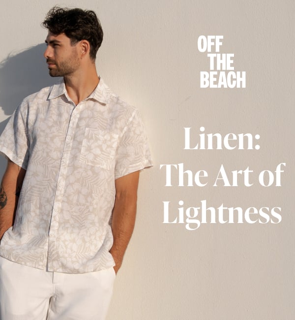 A man leaning against a wall, wearing a floral-patterned linen shirt and white linen pants. 