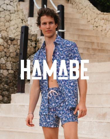 A man descending a staircase, wearing a coordinated set consisting of a floral‑pattern blue shirt and swim shorts from the brand Hamabe. 