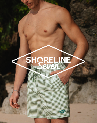 Man on the beach with one hand in the pocket of green‑and‑white check swim shorts from Shoreline Seven. 