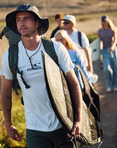 A man walking with a group, holding a surfboard, and wearing a T-shirt and swim trunks from the brand O&rsquo;Neill.