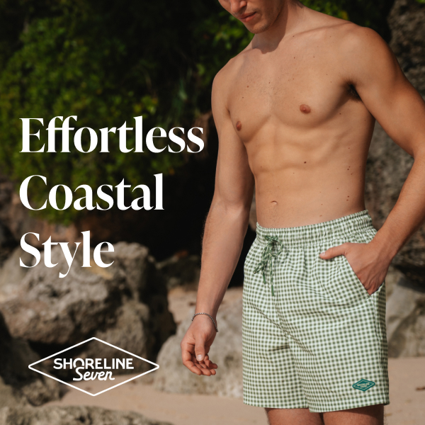 Man on the beach with one hand in the pocket of green-and-white checkered swim trunks by Shoreline Seven.