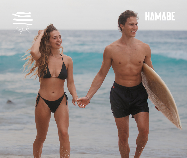 A woman and a man holding hands while walking on the beach, wearing black swimsuits. 