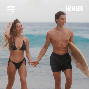 A woman and a man holding hands while walking on the beach, wearing black swimsuits. 
