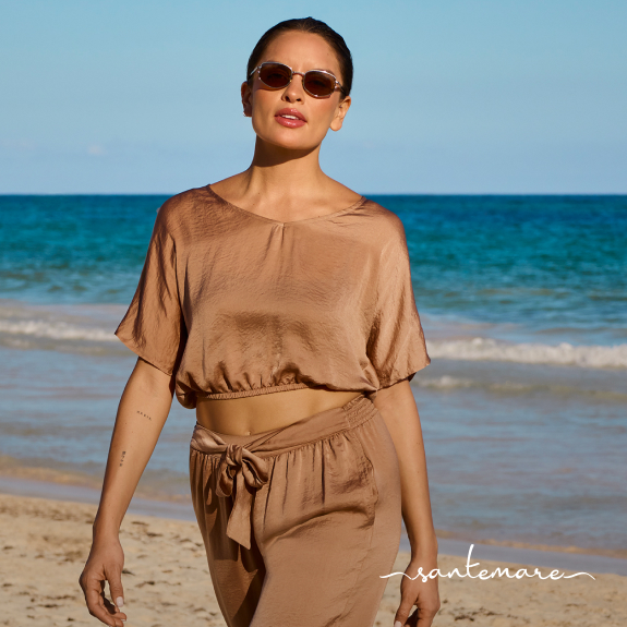 A woman walking along the beach, wearing a coordinated set consisting of a beige satin top and pants from the brand Santemare. 