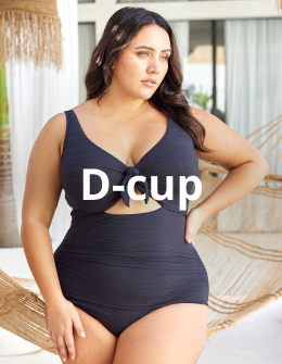 A woman standing in front of a hammock, posing with one hand on her hip, wearing a black D‑cup one-piece swimsuit from the brand Artesands.