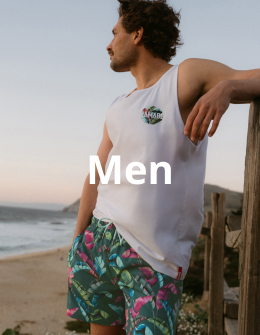 A man near the beach, standing against a fence and holding a surfboard, wearing a tank top and swim trunks from the brand Hamabe.	