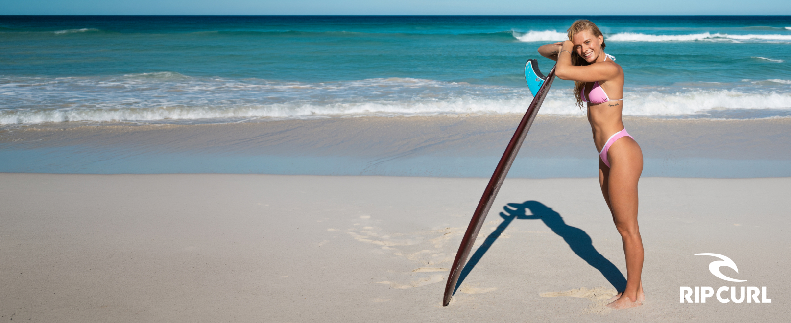 Woman on the beach leaning against a surfboard, wearing a pink and white Rip Curl swimsuit.