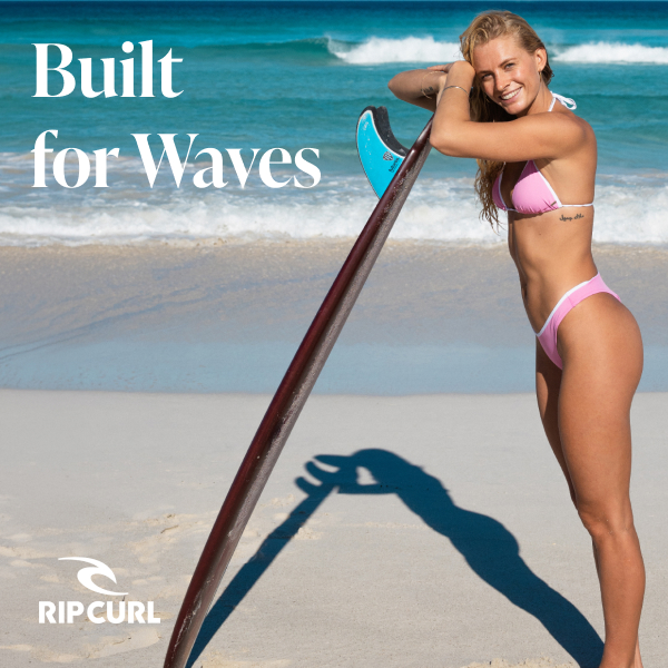 Woman on the beach leaning against a surfboard, wearing a pink and white Rip Curl swimsuit.