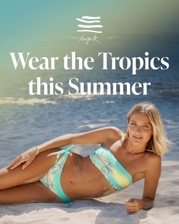 A woman sitting on the beach wearing a bikini set with a tropical landscape pattern from the brand Tropik.