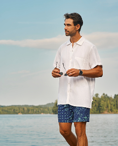 Man walking on the beach wearing a white shirt and blue patterned volley shorts from Shoreline Seven.	