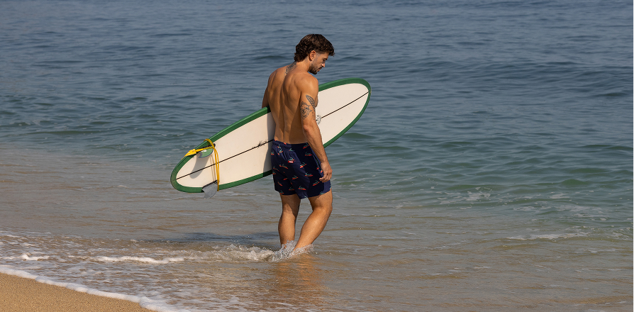 Man holding a surfboard in the water, wearing crocodile-print swim trunks from the Hamabe brand.	