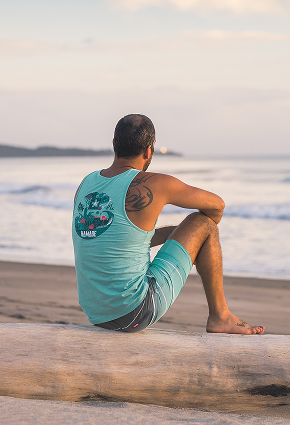 Man sitting on the beach, back to the camera, wearing a blue tank top from Hamabe.
