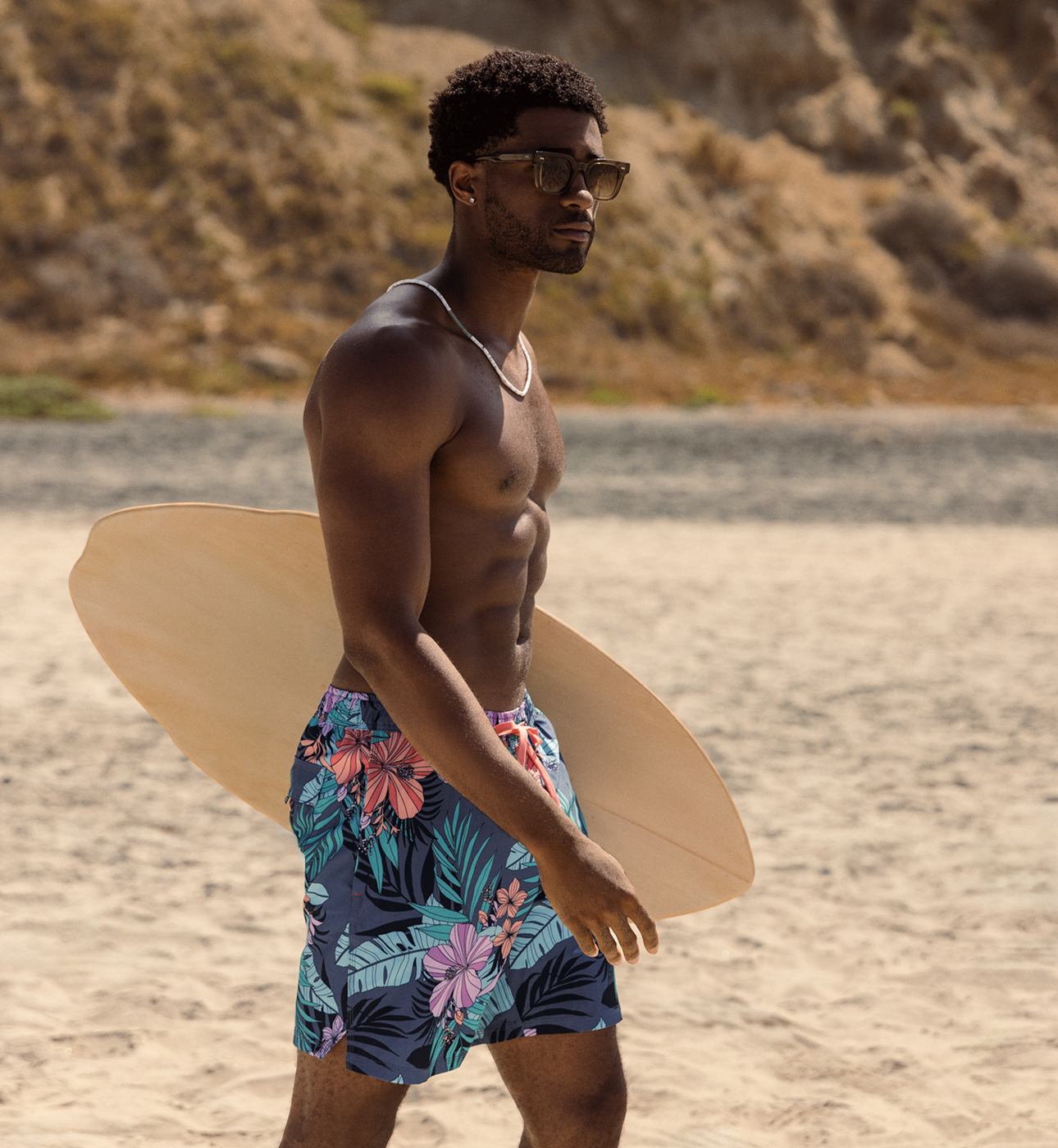 Man walking on the beach holding a surfboard, wearing SAXX swim shorts with an exotic floral print.