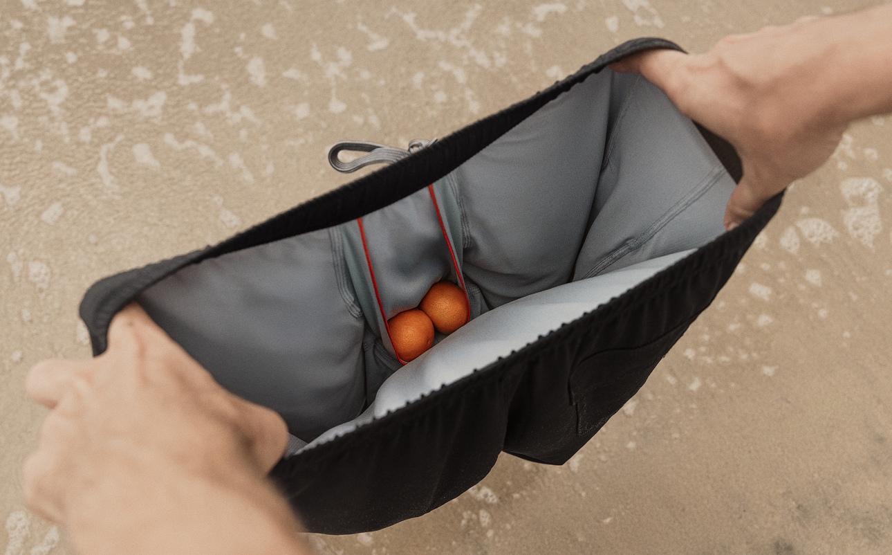 Man holding black SAXX swim shorts, demonstrating the BallPark Pouch using fruit.