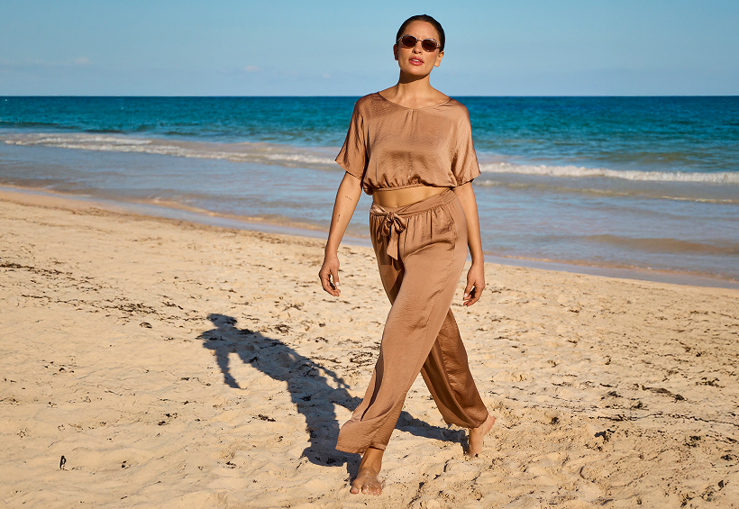 Woman walking on the beach and wearing a beige satin top and pants from the brand Santemare. 