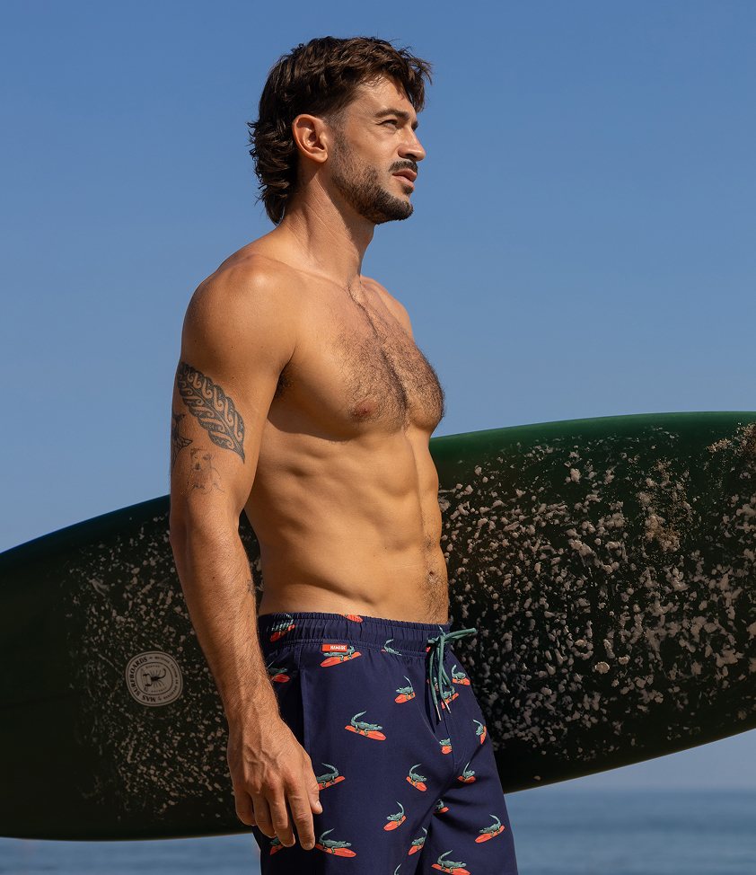 Homme tenant une planche de surf et portant un maillot au motif de crocodiles de la marque Hamabe.